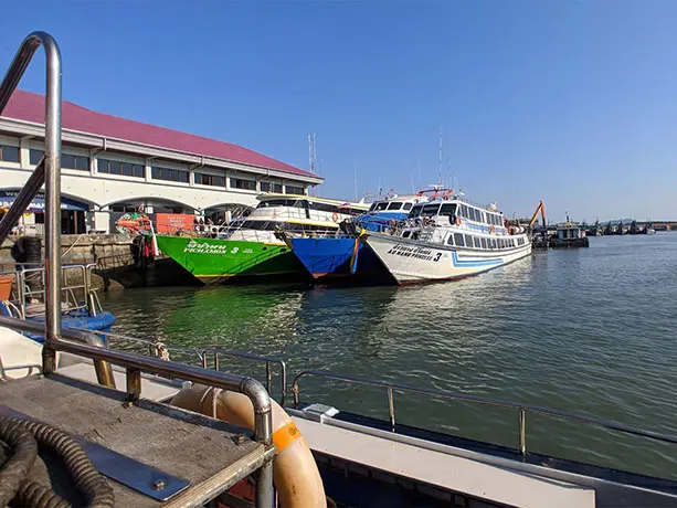 Multiple passenger ferry boats, including a green ferry named Pichamon 3 and a white ferry named Ao Nang Princess 3, docked alongside the main terminal building at Rassada Pier in Phuket